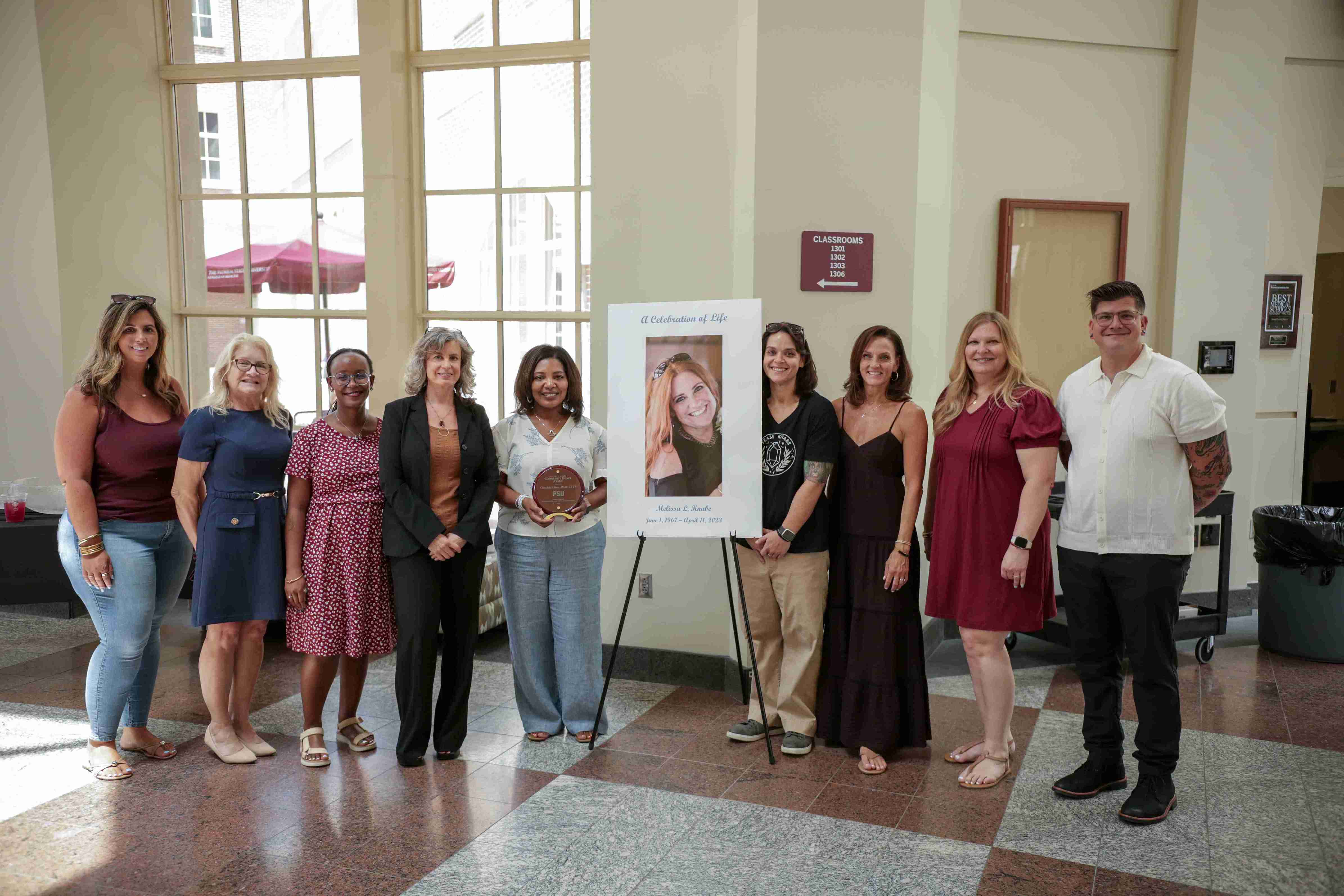 a group of nine people pose together for a photo in a bright atrium around a "A Celebration of Life" memorial poster for Melissa L. Knabe, with one woman in the center holding an award plaque.