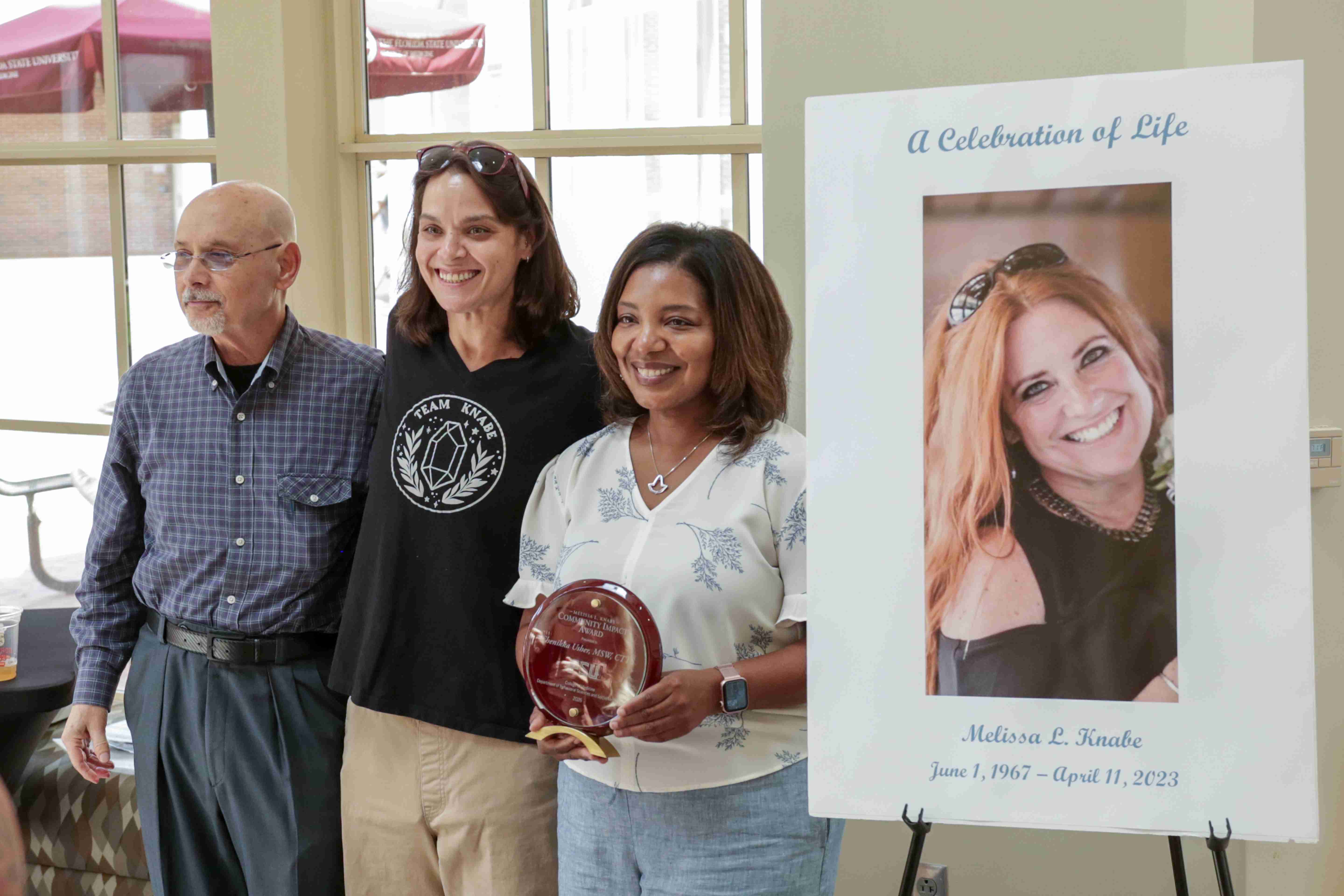 hree people stand smiling next to a memorial poster titled "A Celebration of Life" for Melissa L. Knabe, while the woman in the center proudly displays a round plaque.