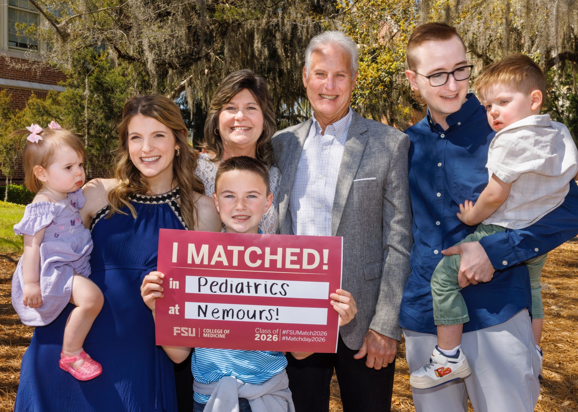 Hope Wolmarans poses with her husband, her two children, her in-laws and her nephew, who is holding her match card that says she matched in pediatrics at Nemours Children's Hospital Florida.