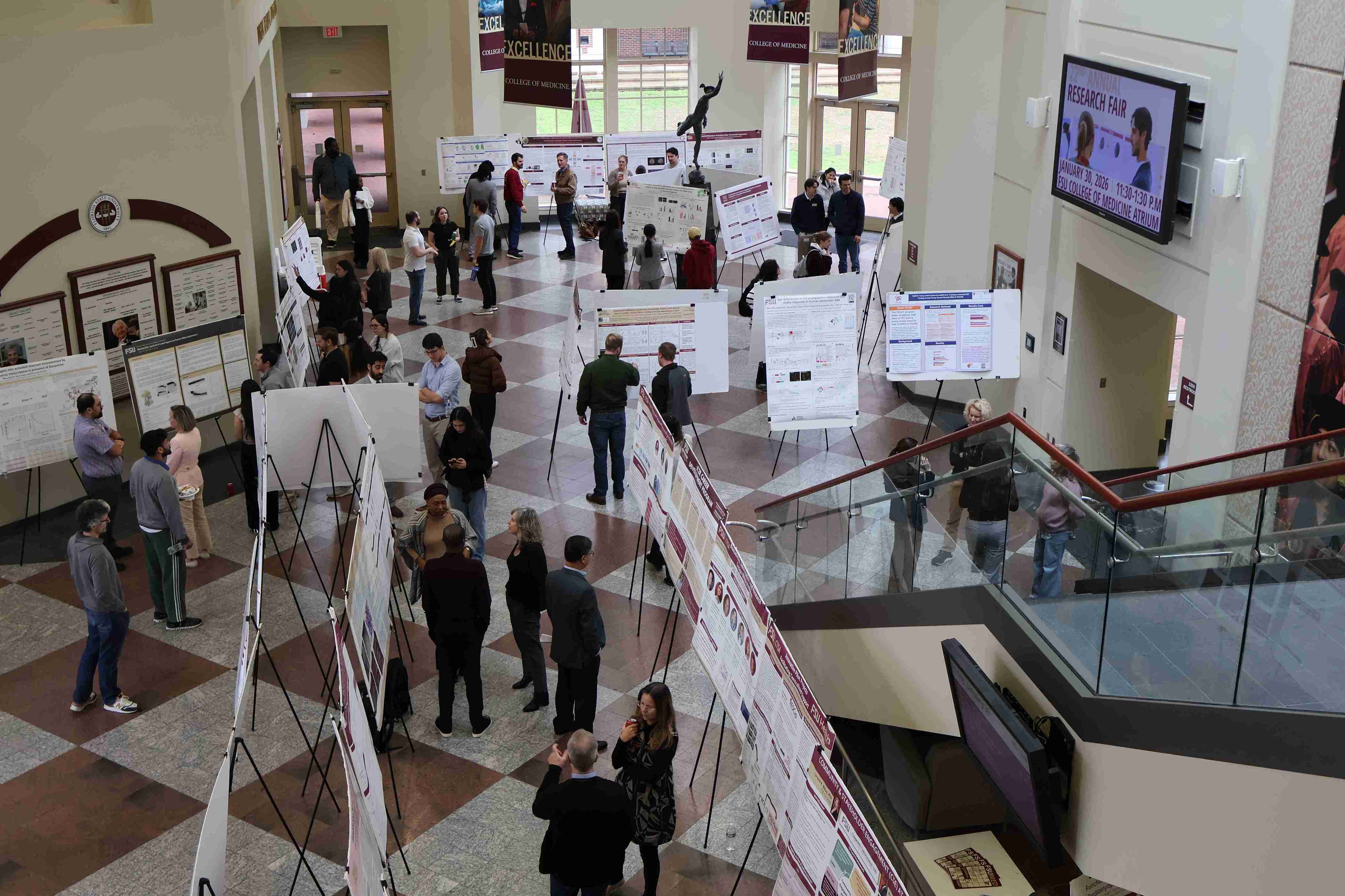 Upper‑floor view overlooking a research poster event, with attendees gathered below reviewing and discussing posters.