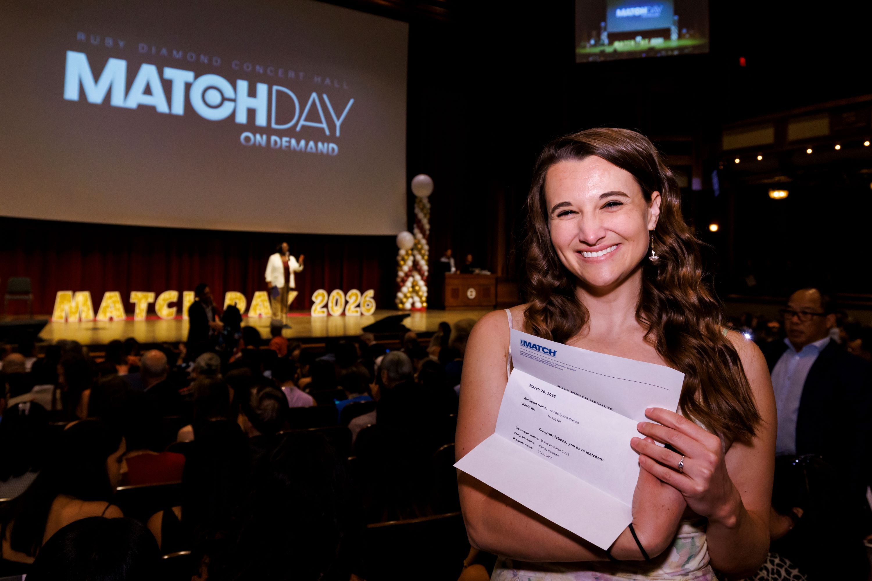 Kimberly Keenan beams as she shows off her letter saying she matched in family medicine at Ascension St. Vincent Medical Center in Jacksonville. In the background, College of Medicine Dean Alma B. Littles, M.D., speaks from the stage.