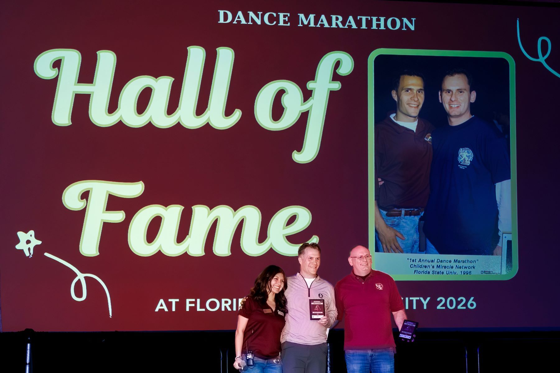 Nicole Lang of the DM@FSU Executive Board, poses with co-founders Bryan Seaquist, center, and Travis Ferguson after they were named to the DM@FSU Hall of Fame. The slide in the background includes a photo of them at the first Dance Marathon at FSU, 31 years ago.