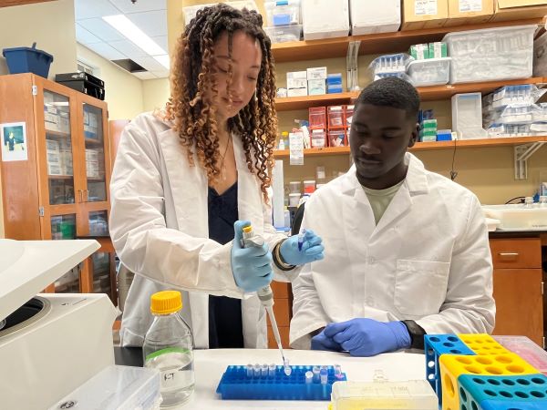 Image shows two dark-skinned scientists in a research lab. The female is standing and pipetting. The male is seated and observing. Both are wearing white lab coats. 