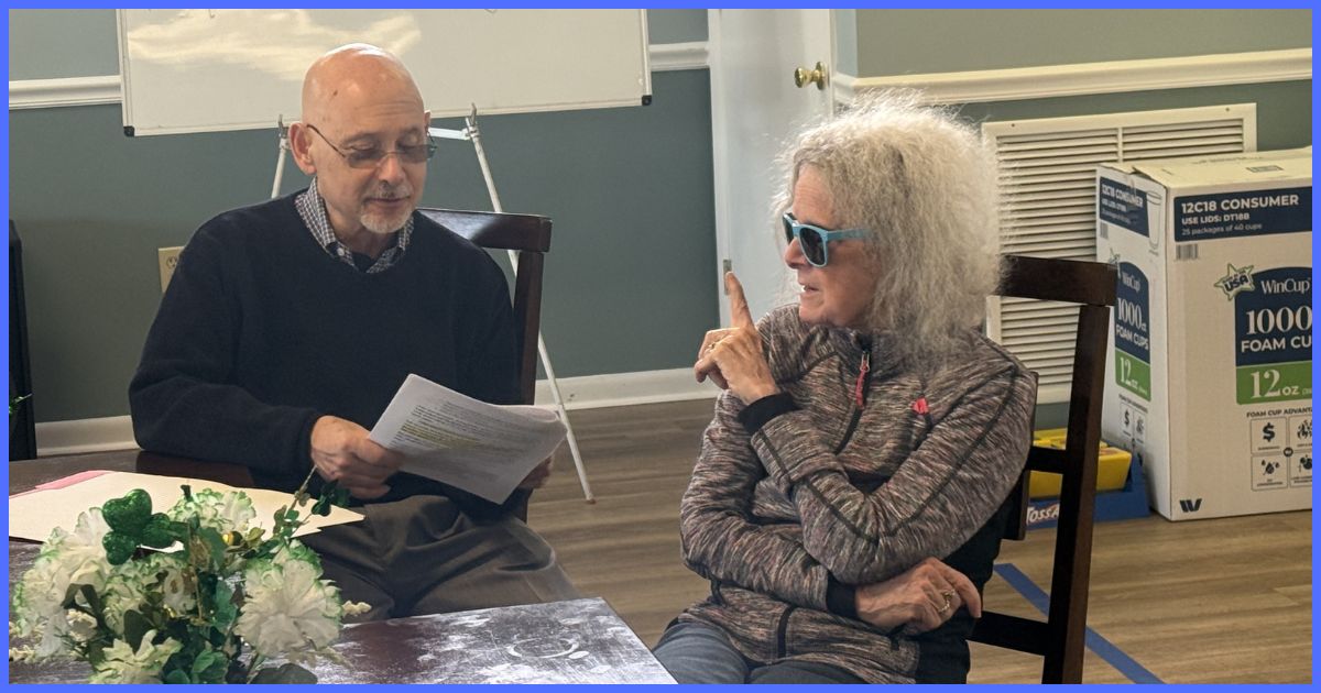 A man reviews a printed document with an older woman seated beside him as they engage in conversation during a small group meeting in a community room.