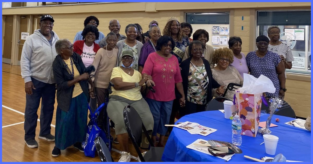 A group of older adults pose together and smile for a photo during a community gathering at an indoor recreation center, with tables, informational materials, and decorations visible in the foreground.