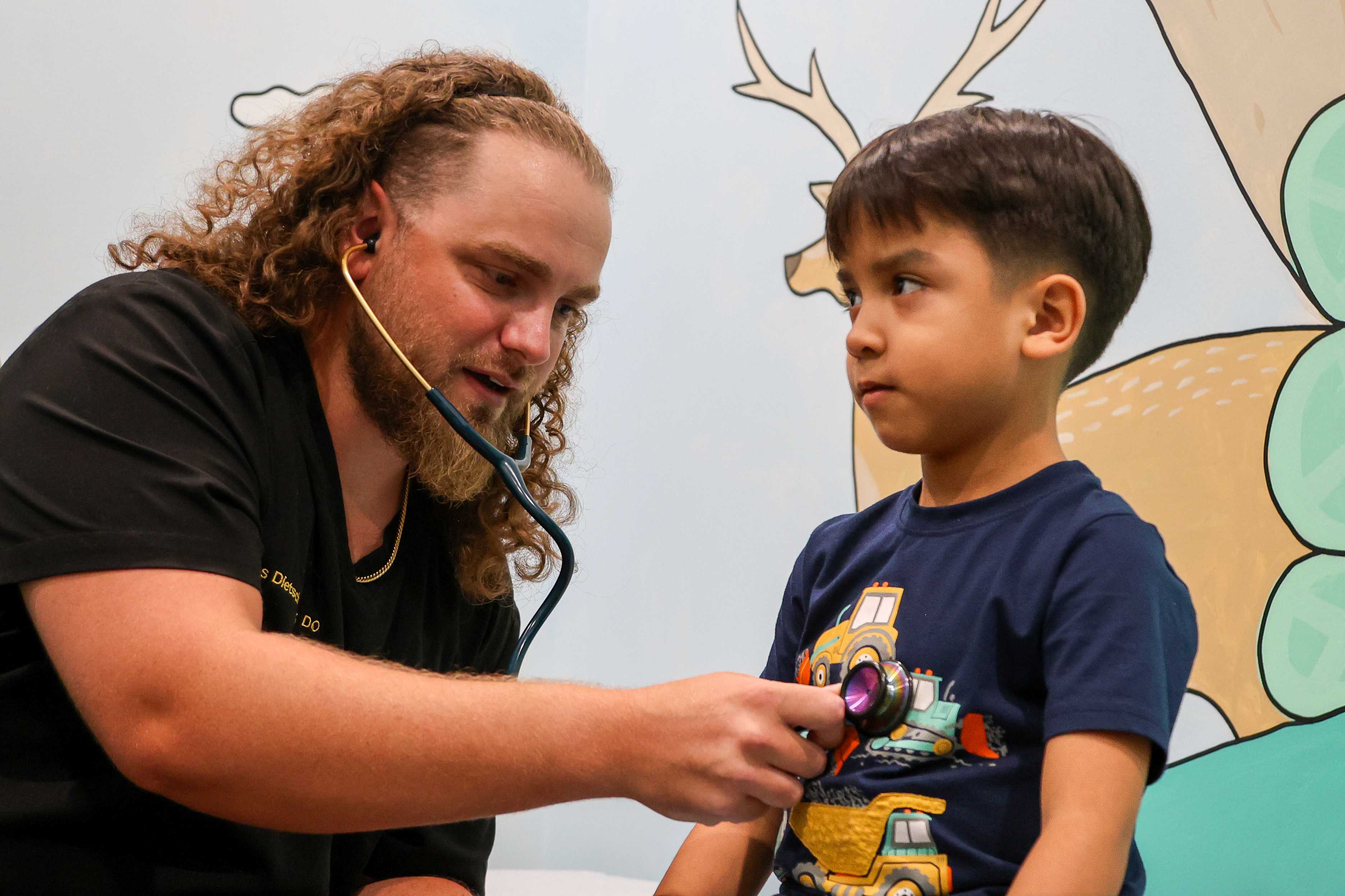 A male physician resident with a stethoscope listens to the heart of a young boy who is sitting on an exam table. 