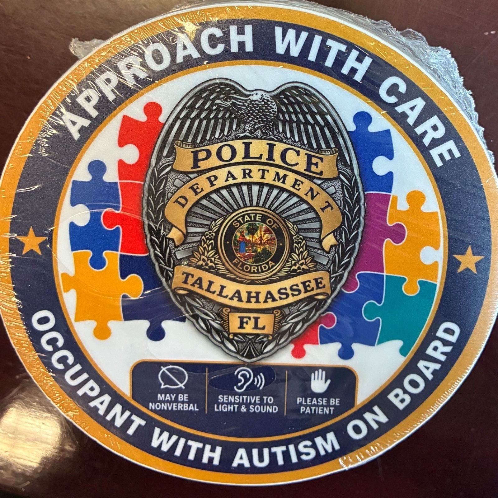 Photo of sticker for auto back window saying "Approach with Care" and "Occupant with Autism on Board" circling a Tallahassee Police Department badge.