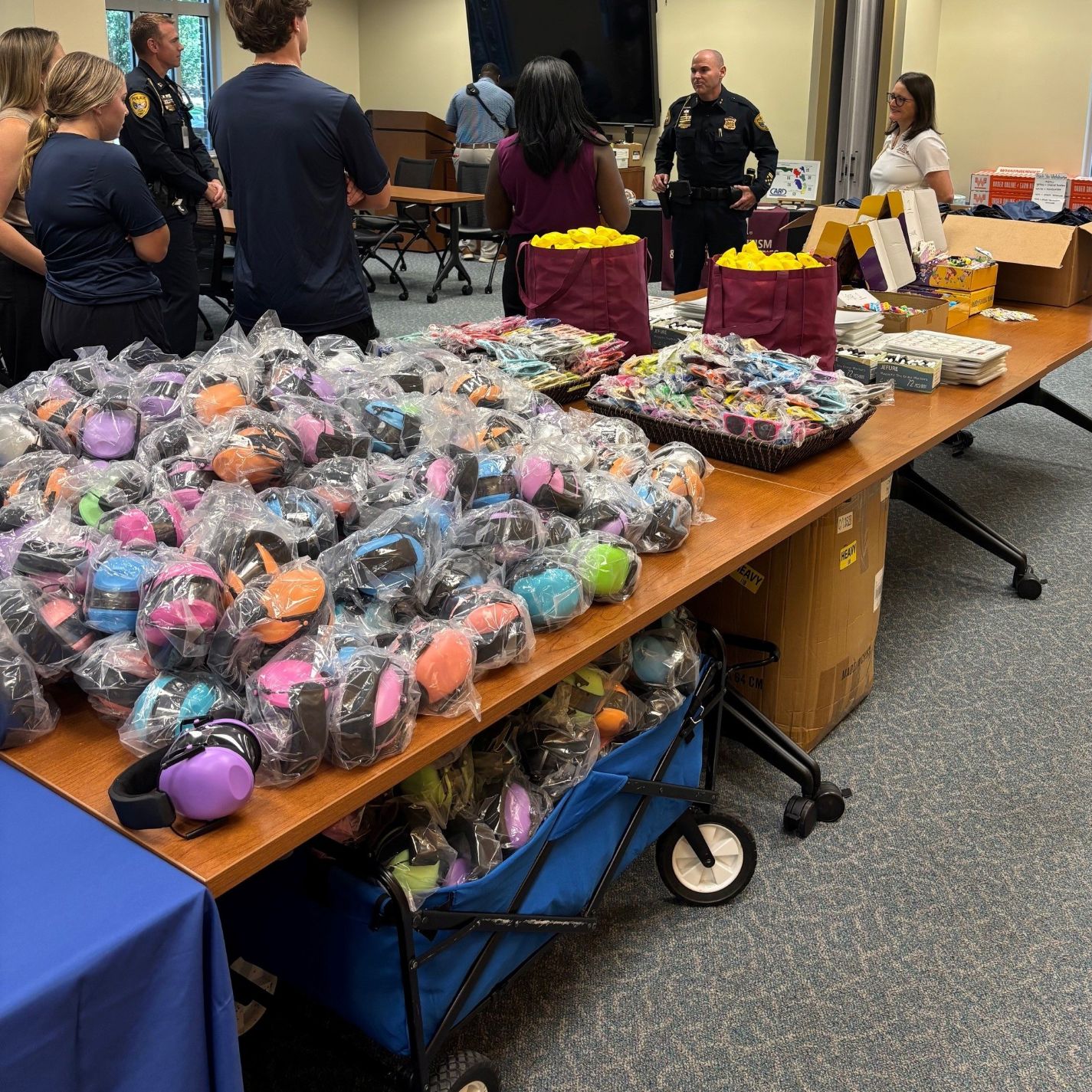 A man and women speak to a group of people standing next to a table of sensory comfort items to be stuffed into bags.